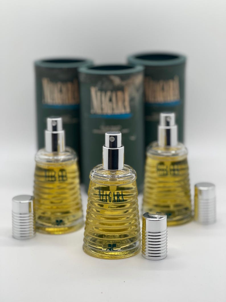 May include: Three glass perfume bottles with silver sprayers and caps, filled with yellow liquid. The bottles are labeled "Migara" and are accompanied by three green cylindrical boxes with the same label. The bottles have a ribbed design.