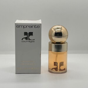 May include: A perfume bottle and box set. The bottle is cylindrical with a gold cap and a gold band. The box is rectangular and gray with the word "empreinte" and the brand logo printed on it. The perfume inside is a light amber color.