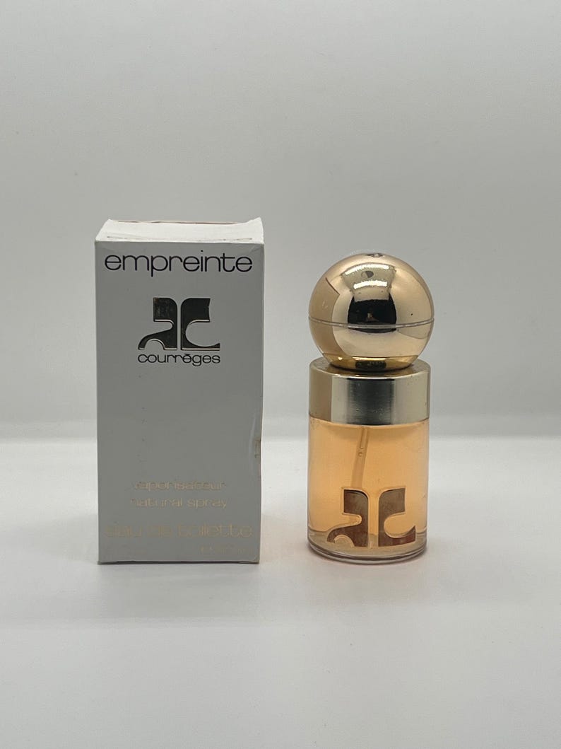 May include: A perfume bottle and box set. The bottle is cylindrical with a gold cap and a gold band. The box is rectangular and gray with the word "empreinte" and the brand logo printed on it. The perfume inside is a light amber color.