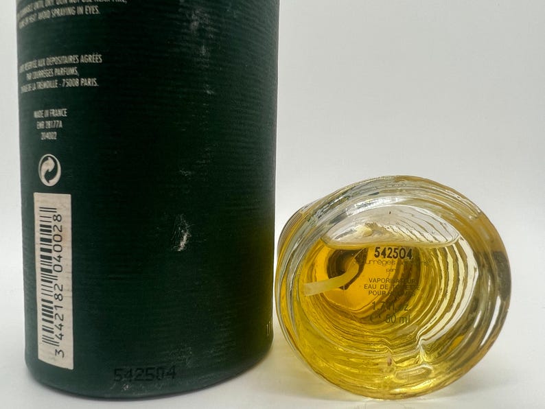 May include: A perfume bottle with a clear, textured glass base and a dark green cylindrical container. The bottle contains a yellow liquid. The text "542504" is visible on the bottle and container. The bottle is labeled "VAPORISATEUR EAU DE PARFUM" and contains 60 ml.
