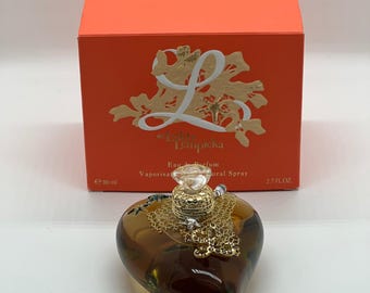 May include: A heart-shaped amber glass perfume bottle with a gold-toned honeycomb-style cap and a clear crystal-like topper, positioned in front of an orange box labeled L de Lolita Lempicka, 80 ml, 2.7 fl. oz. Eau de Parfum natural spray.