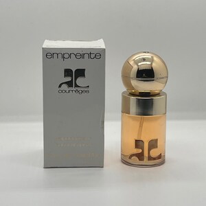 May include: A perfume bottle and box set. The bottle has a gold spherical cap and a clear body with a gold logo. The box is white with the word "empreinte" and a logo in black. The perfume is a light amber color.