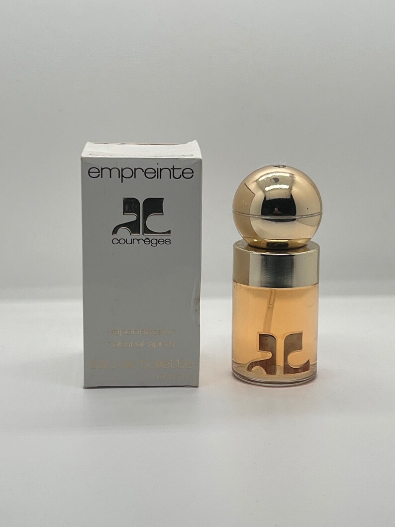 May include: A perfume bottle and box set. The bottle has a gold spherical cap and a clear body with a gold logo. The box is white with the word "empreinte" and a logo in black. The perfume is a light amber color.