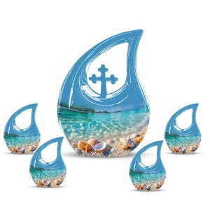 May include: Five blue teardrop-shaped ornaments with a beach scene inside. The largest ornament features a cross design. The beach scene includes clear turquoise water, sand, and seashells. The ornaments are set against a white background.