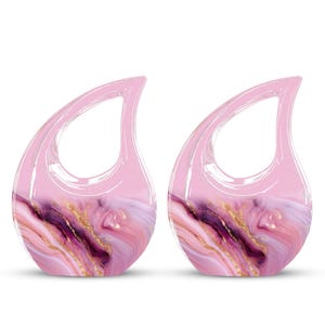 May include: Two pink teardrop-shaped decorative items with a marbled design. The marble pattern features shades of pink, purple, and gold. The top portion of each item is a solid, translucent pink, and they have a loop at the top.