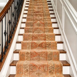Puede incluir: Una alfombra larga y estrecha para escaleras con un patrón geométrico repetitivo en tonos naranja quemado, terracota y beige apagado. La alfombra tiene una apariencia tejida y texturizada con bordes decorativos sobre una escalera de madera.