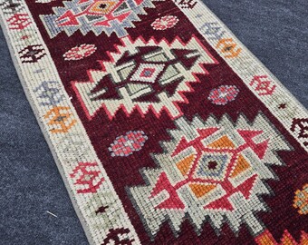 Op de afbeelding: Een close-up van een kleurrijk, geometrisch patroon tapijt met een bordeauxrode achtergrond. Het tapijt heeft een witte rand met ingewikkelde ontwerpen in tinten rood, oranje, blauw en grijs.