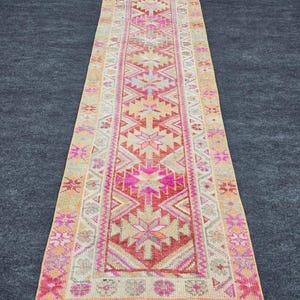 May include: A rectangular, vintage-style runner rug with a central geometric pattern in shades of pink, orange, and beige. The rug features a border with floral and geometric motifs. The rug is approximately 305 cm long.