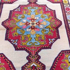 May include: A vintage rug with a cream background and a central medallion in shades of blue, red, yellow, and pink. The medallion features intricate geometric patterns and floral motifs. The rug has a red border with a repeating pattern.
