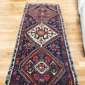 May include: A long, narrow rug with a traditional Persian design. The rug has a dark blue background with intricate patterns in shades of red, orange, pink, and white. The rug has a fringe border.