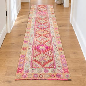 Puede incluir: Una alfombra de pasillo larga y estrecha con un patrón geométrico vibrante en tonos rosa intenso, naranja, beige y azul claro. El diseño consta de motivos repetitivos en forma de estrella y bordes intrincados, creando una pieza textil simétrica.