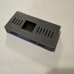 May include: A gray rectangular electronic device case with ventilation slots. The case has a rectangular opening in the center and several rectangular openings on the sides. The case is made of a matte material.
