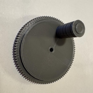 May include: A gray plastic gear with a round, toothed edge and a smaller, round, smooth center. A gray plastic handle is attached to the gear.