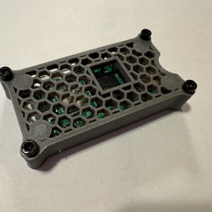 May include: A gray 3D printed case with a honeycomb pattern and four black screws. The case has a cutout for a circuit board.