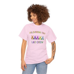Może przedstawiać: Różowy T-shirt z napisem "Delivering Tiny Peeps L&D Crew" i rzędem pięciu kolorowych królików z literami "P-E-E-P-S" na brzuchu.
