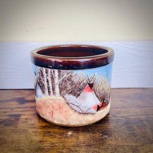 May include: A brown ceramic jar with a painted scene of teepees in a forest. The jar has a brown rim and a light brown base.