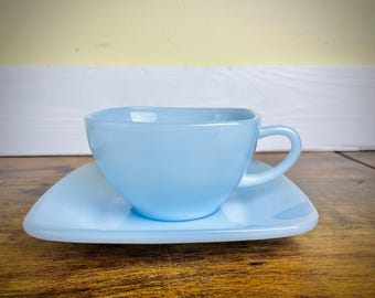 May include: A light blue teacup and saucer set. The cup has a square shape with a rounded handle, and the saucer is a matching light blue color. The set is made of ceramic or glass and is placed on a wooden surface.