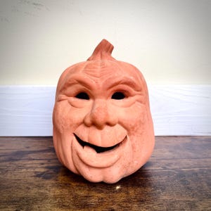 May include: A terracotta-colored pumpkin face with a smiling expression. The pumpkin has a textured surface, black eye sockets, and a stem. This decorative piece is suitable for Halloween or fall decor.