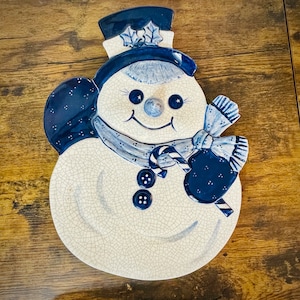 May include: A ceramic snowman plate with a blue and white glaze. The snowman has a blue hat, scarf, and mittens. The plate is perfect for serving holiday snacks or desserts.
