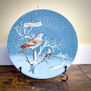 May include: A decorative plate with a winter scene featuring two birds perched on a bare tree. The plate has a blue background with white snowflakes and a banner that reads "Noël 1971". The birds have brown, white, and blue feathers.