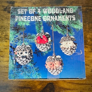 May include: A box of four woodland pinecone ornaments. The ornaments are decorated with a cardinal, two bunnies, and two squirrels. The box is blue with white lettering that reads "Set of 4 Woodland Pinecone Ornaments".