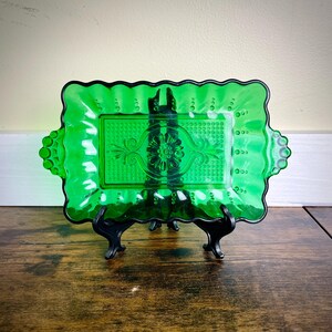 May include: A rectangular, emerald green glass tray with a decorative floral and scroll design. The tray has a scalloped edge and is supported by a black metal stand. The tray is approximately 10 inches long and 6 inches wide.