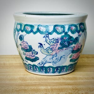 May include: A white ceramic planter with a wide opening and a turquoise interior. The exterior features hand-painted designs of birds, flowers, and foliage in shades of blue, pink, and green. The planter sits on a wooden surface.