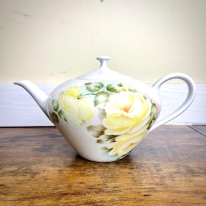 May include: White ceramic teapot with a lid and handle. The teapot is decorated with a large yellow rose design with green leaves and pink accents. The teapot is sitting on a wooden surface.