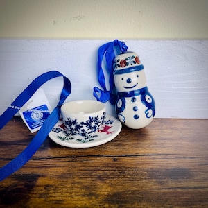 May include: A ceramic snowman ornament with a blue ribbon, a small teacup, and a saucer. The snowman has a blue scarf and hat with red floral accents. The teacup and saucer feature a blue and white floral design.