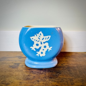 May include: A light blue ceramic planter with a white floral design. The planter has a rounded base and a square top.