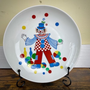 May include: White ceramic plate featuring a colorful clown design. The clown has a checkered vest, red pants, and a blue hat. The plate is decorated with colorful balls and dots, and is displayed on a black metal stand.