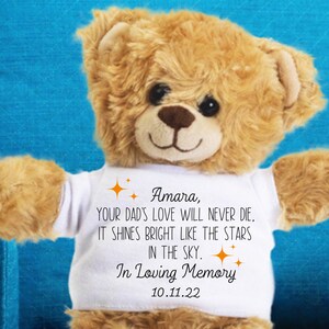 May include: A brown teddy bear wearing a white t-shirt with the text "Amara, YOUR DAD'S LOVE WILL NEVER DIE. IT SHINES BRIGHT LIKE THE STARS IN THE SKY. In Loving Memory 10.11.22"