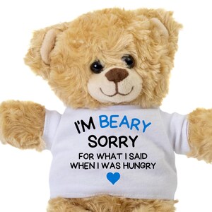 May include: A plush, tan teddy bear wearing a white t-shirt. The shirt has the text "I'M BEARY SORRY FOR WHAT I SAID WHEN I WAS HUNGRY" in black and blue, with a blue heart below. The bear has black eyes and a brown nose.