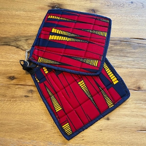 Peut inclure: Deux maniques matelassées avec un tissu rouge vif orné d'un motif géométrique jaune et noir. Les maniques ont une bordure bleu marine et une boucle pour les suspendre. Le design est symétrique et attrayant.