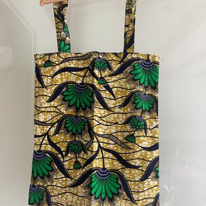 Op de afbeelding: Een gele, groene en blauwe tote bag met een bloemenpatroon. De tas heeft twee lange hengsels.