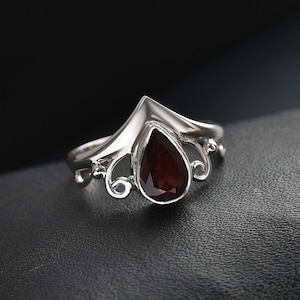 May include: A silver ring with a pear-shaped, dark red gemstone. The ring has a decorative, curved design around the gemstone and on the sides. The ring is set against a dark background.