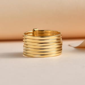 May include: A gold ring with a multi-layered design. The ring has seven horizontal bands, giving a stacked look. The ring is made of a shiny, reflective metal, set against a neutral background.