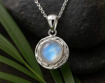 May include: A silver pendant necklace featuring a round, iridescent moonstone cabochon. The moonstone is set in a textured, circular frame and hangs from a delicate chain. The pendant is displayed against a dark background with green foliage.