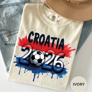 Może przedstawiać: Kremowa koszulka z czarnym napisem "CROATIA" nad liczbami "2026" i piłką nożną. Projekt wykorzystuje elementy czerwone, białe i niebieskie z efektami rozprysk&oacute;w farby. Koszulka leży na niebieskich dżinsach.