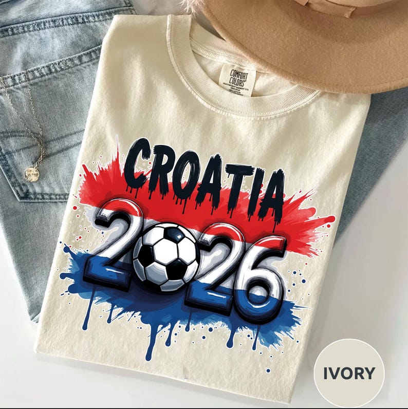 Może przedstawiać: Kremowa koszulka z czarnym napisem "CROATIA" nad liczbami "2026" i piłką nożną. Projekt wykorzystuje elementy czerwone, białe i niebieskie z efektami rozprysk&oacute;w farby. Koszulka leży na niebieskich dżinsach.
