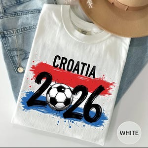 May include: White t-shirt with the word "CROATIA" in black, a soccer ball, and the number "2026". The design features red and blue brushstrokes, with blue paint splatters. The shirt is folded on top of blue jeans.