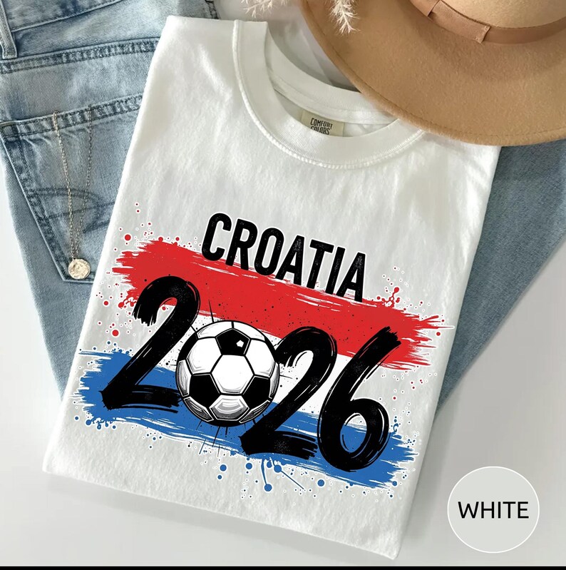 May include: White t-shirt with the word "CROATIA" in black, a soccer ball, and the number "2026". The design features red and blue brushstrokes, with blue paint splatters. The shirt is folded on top of blue jeans.