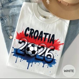 Może przedstawiać: Biała koszulka z napisem "CROATIA" w kolorze czarnym, nad liczbami "2026" i piłką nożną. Projekt wykorzystuje kolory czerwony, biały i niebieski z efektem rozprysku. Koszulka jest złożona na niebieskich dżinsach.
