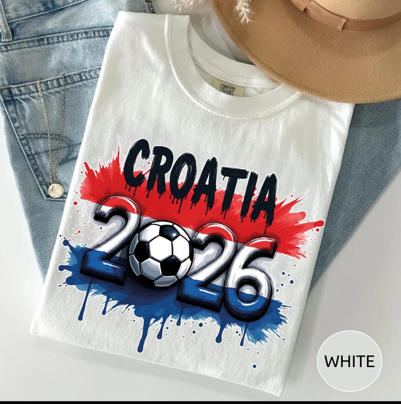Może przedstawiać: Biała koszulka z napisem "CROATIA" w kolorze czarnym, nad liczbami "2026" i piłką nożną. Projekt wykorzystuje kolory czerwony, biały i niebieski z efektem rozprysku. Koszulka jest złożona na niebieskich dżinsach.