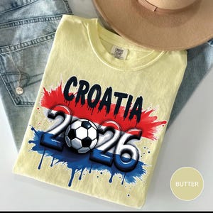 Może przedstawiać: Jasnoż&oacute;łta koszulka z czarnym napisem "CROATIA", nad liczbami "2026" i piłką nożną. Projekt zawiera kolory czerwony, biały i niebieski, z efektem rozprysku. Beżowy kapelusz i niebieskie dżinsy są w tle.