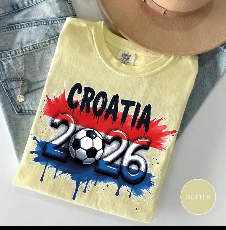 Może przedstawiać: Jasnoż&oacute;łta koszulka z czarnym napisem "CROATIA", nad liczbami "2026" i piłką nożną. Projekt zawiera kolory czerwony, biały i niebieski, z efektem rozprysku. Beżowy kapelusz i niebieskie dżinsy są w tle.