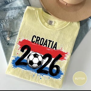 May include: A light yellow t-shirt with the word "CROATIA" in black, above the numbers "2026" and a soccer ball graphic. The design incorporates red, white, and blue brushstrokes, and the shirt is folded on a pair of blue jeans.