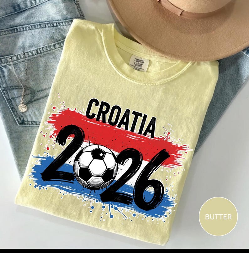 May include: A light yellow t-shirt with the word "CROATIA" in black, above the numbers "2026" and a soccer ball graphic. The design incorporates red, white, and blue brushstrokes, and the shirt is folded on a pair of blue jeans.