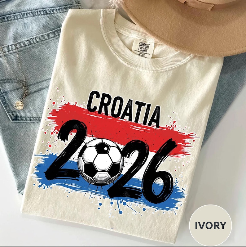 May include: Ivory-colored t-shirt with the word "CROATIA" in black, above the number "2026" and a soccer ball graphic. The design features red and blue brushstrokes, with blue paint splatters. The shirt is part of a sports-themed apparel collection.