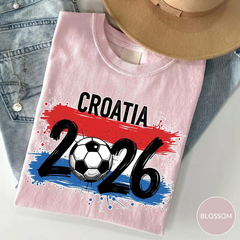 May include: A light pink t-shirt featuring the word "CROATIA" in black above the number "2026". The number "2026" is in black with a soccer ball in the center. The design includes red and blue brushstrokes.